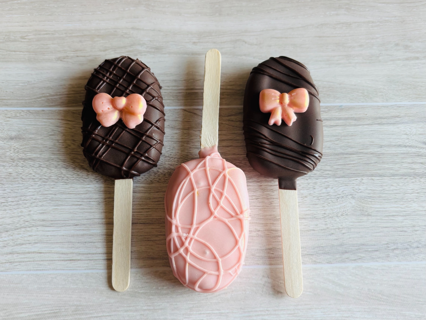 Three cakecicles in dark chocolate and pink with hand-piped bow decorations